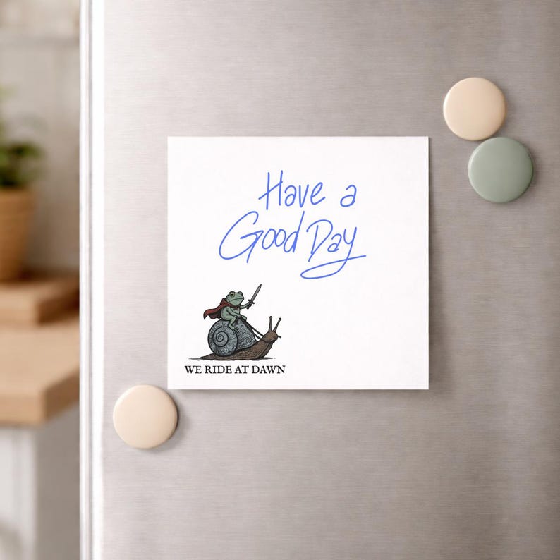 May include: White square magnet with the text "Have a Good Day" in blue script. A cartoon frog in a cape and holding a sword rides a snail. The text "WE RIDE AT DAWN" is below the illustration. Beige and green magnets are on the right.