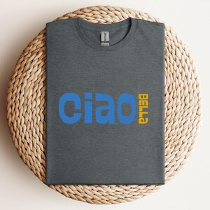 May include: A dark gray t-shirt with the words "ciao BELLA" printed in blue and gold. The shirt is folded and placed on a woven, circular surface.