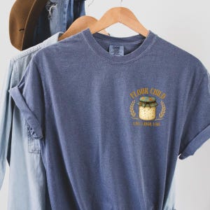 May include: A blue t-shirt featuring the text "FLOUR CHILD" above a bread jar illustration, with "LIVE. LAUGH. LOAF." underneath. The shirt is displayed on a wooden hanger, alongside other clothing items.