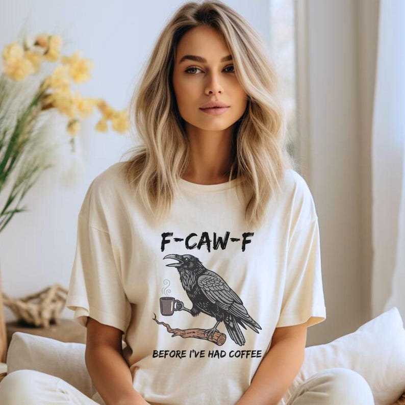 May include: Cream-colored t-shirt featuring a black raven perched on a branch, holding a coffee cup. The text "F-CAW-F" is above the raven, with "BEFORE I'VE HAD COFFEE" below. The shirt has short sleeves.