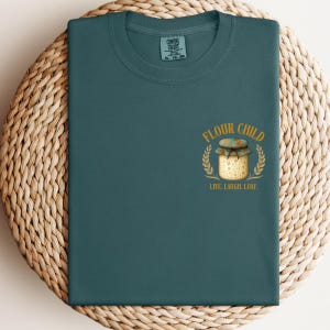 May include: Teal crew neck t-shirt with a graphic. The graphic shows a jar with the words "FLOUR CHILD" and "LIVE. LAUGH. LOAF.". The jar is surrounded by a wheat-like design. The shirt is a solid teal colour.