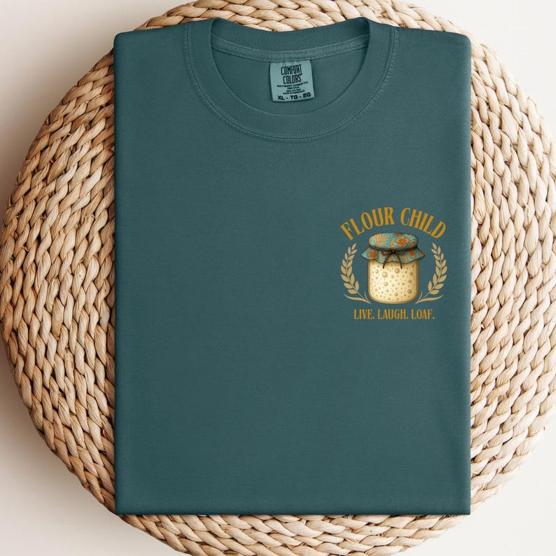 May include: Teal crew neck t-shirt with a graphic. The graphic shows a jar with the words "FLOUR CHILD" and "LIVE. LAUGH. LOAF.". The jar is surrounded by a wheat-like design. The shirt is a solid teal colour.