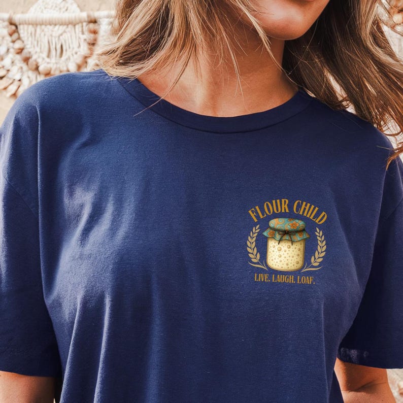 May include: A navy blue t-shirt with a graphic. The graphic includes the words "Flour Child" above a jar illustration with a lid, framed by wheat stalks. The text "Live, Laugh, Loaf" is below.