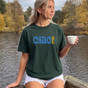 May include: Dark green t-shirt with the word "Ciao" in blue and "Bella" in yellow. The shirt is worn by a person holding a white cup. The person is sitting on a wooden dock near a body of water.