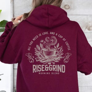 May include: Burgundy hoodie with a coffee-themed graphic on the back. The design includes a coffee cup, steam, and the text "ALL YOU NEED IS LOVE, AND A CUP OF COFFEE" and "RISE&GRIND MORNING BLEND".