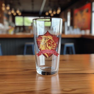 May include: A clear glass pint glass with a red and gold Gryffindor crest featuring a lion. The crest has the words "Gryffindor" in gold lettering.