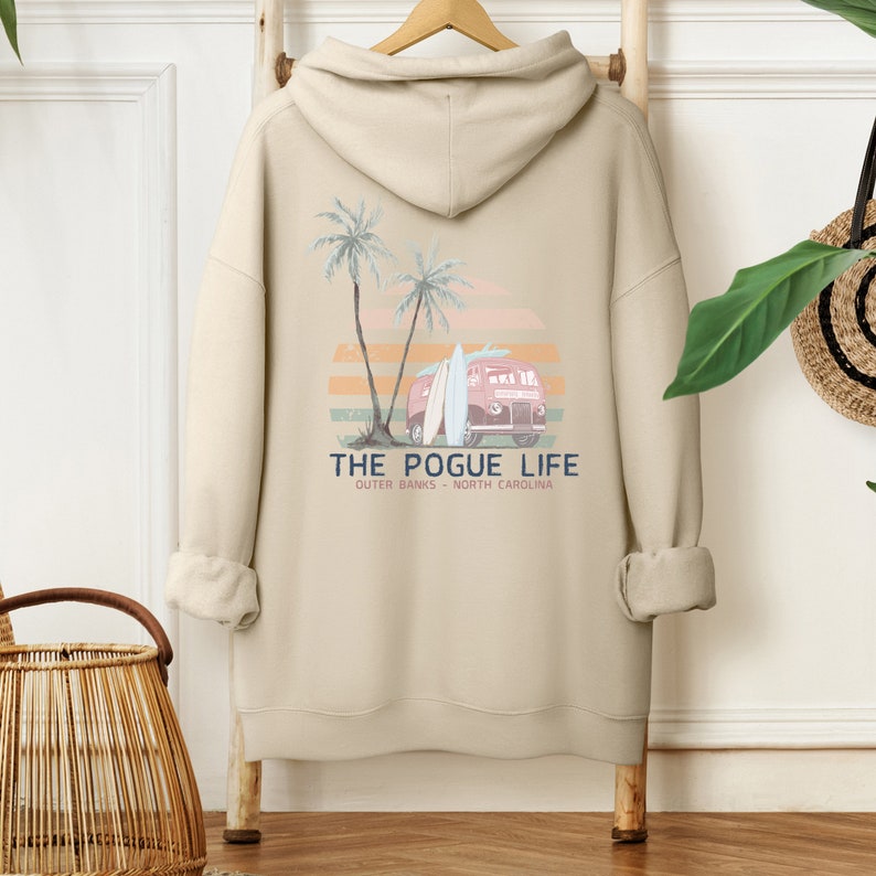 Op de afbeelding: Beige hoodie met een grafische print van een vintage busje met surfplanken en palmbomen. De tekst "THE POGUE LIFE" is onder de afbeelding gedrukt, met daaronder de tekst "OUTER BANKS - NORTH CAROLINA".