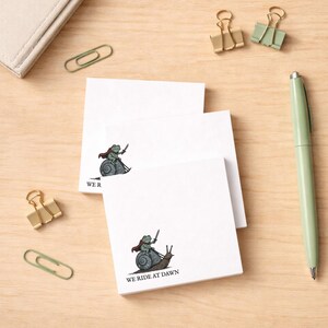May include: White square sticky notes featuring a cartoon frog warrior riding a snail, with a sword and cape. The text "WE RIDE AT DAWN" is printed below the illustration. Stationery items, including paperclips and a pen, are also visible.