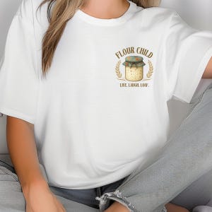 May include: White t-shirt with a graphic of a jar with a floral lid, wheat stalks, and the text "FLOUR CHILD. LIVE. LAUGH. LOAF." The design uses brown, green, and beige colours. A casual, comfortable garment.