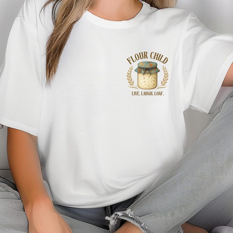 May include: White t-shirt with a graphic of a jar with a floral lid, wheat stalks, and the text "FLOUR CHILD. LIVE. LAUGH. LOAF." The design uses brown, green, and beige colours. A casual, comfortable garment.