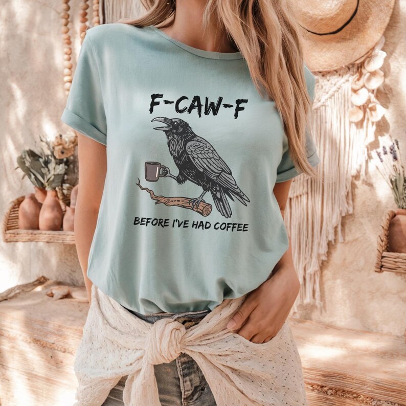 May include: A light blue t-shirt with the text "F-CAW-F" above a black raven holding a coffee cup, perched on a branch. Below the raven is the text "BEFORE I'VE HAD COFFEE". The shirt has rolled-up sleeves.
