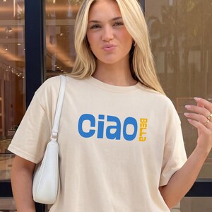 May include: Beige t-shirt with the word "Ciao" in blue and "Bella" in yellow. The shirt is a neutral color and has a relaxed fit. The model is wearing a white shoulder bag.