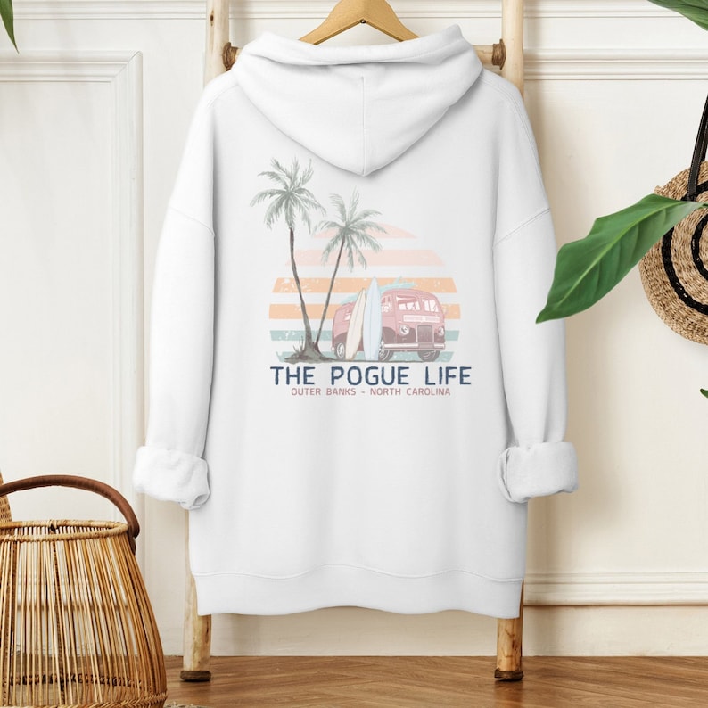 Op de afbeelding: Witte hoodie met een grafische print van een rode vintage bus met surfplanken, palmbomen en een zonsondergang. De tekst "THE POGUE LIFE" is onder de print gedrukt, met daaronder de woorden "OUTER BANKS - NORTH CAROLINA".