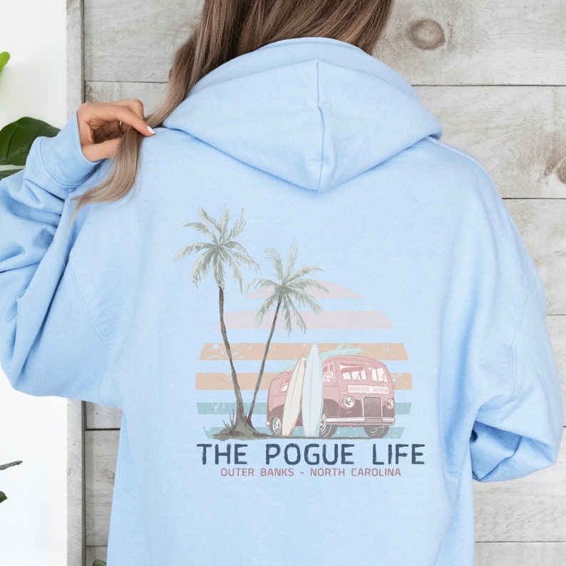 Op de afbeelding: Een lichtblauwe hoodie met een grafische print van een vintage busje met surfplanken, palmbomen en een zonsondergang. De tekst "THE POGUE LIFE" is onder de print gedrukt, met daaronder de woorden "OUTER BANKS - NORTH CAROLINA".