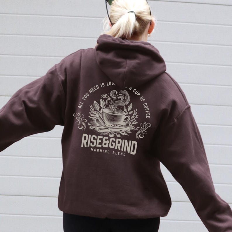 May include: A dark brown hoodie with a coffee-themed graphic on the back. The design includes a coffee cup, leaves, and the text "ALL YOU NEED IS LOV. A CUP OF COFFEE RISE&GRIND MORNING BLEND". The hoodie has a hood and long sleeves.