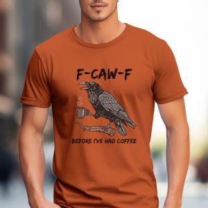 May include: Orange t-shirt with a graphic of a raven holding a coffee cup on a branch. The text "F-CAW-F" is above the raven, and "BEFORE I'VE HAD COFFEE" is below. The raven is black and white.