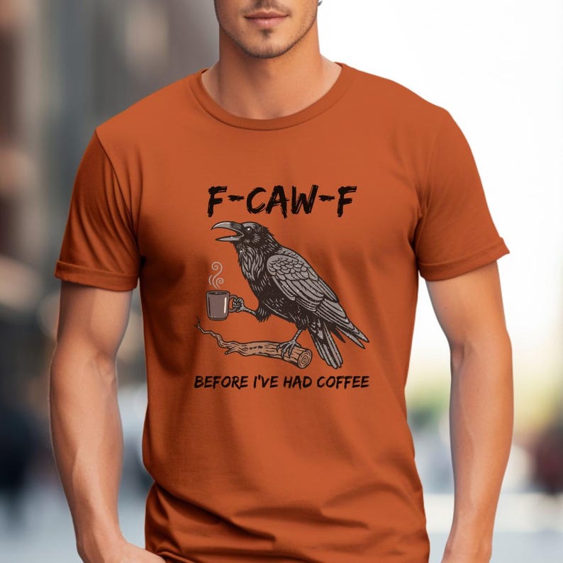 May include: Orange t-shirt with a graphic of a raven holding a coffee cup on a branch. The text "F-CAW-F" is above the raven, and "BEFORE I'VE HAD COFFEE" is below. The raven is black and white.