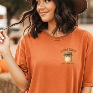 May include: Orange t-shirt featuring the text "Flour Child" and a bread jar illustration. The shirt has a relaxed fit and short sleeves. The text "Live. Laugh. Loaf." is below the jar illustration.