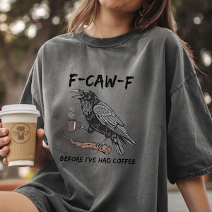 May include: Dark gray t-shirt with a graphic of a raven holding a coffee cup, perched on a branch. Text above reads "F-CAW-F" and below "BEFORE I'VE HAD COFFEE". A person is holding a coffee cup.