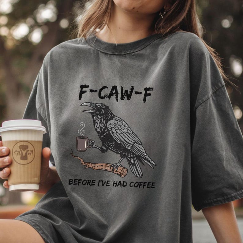 May include: Dark gray t-shirt with a graphic of a raven holding a coffee cup, perched on a branch. Text above reads "F-CAW-F" and below "BEFORE I'VE HAD COFFEE". A person is holding a coffee cup.