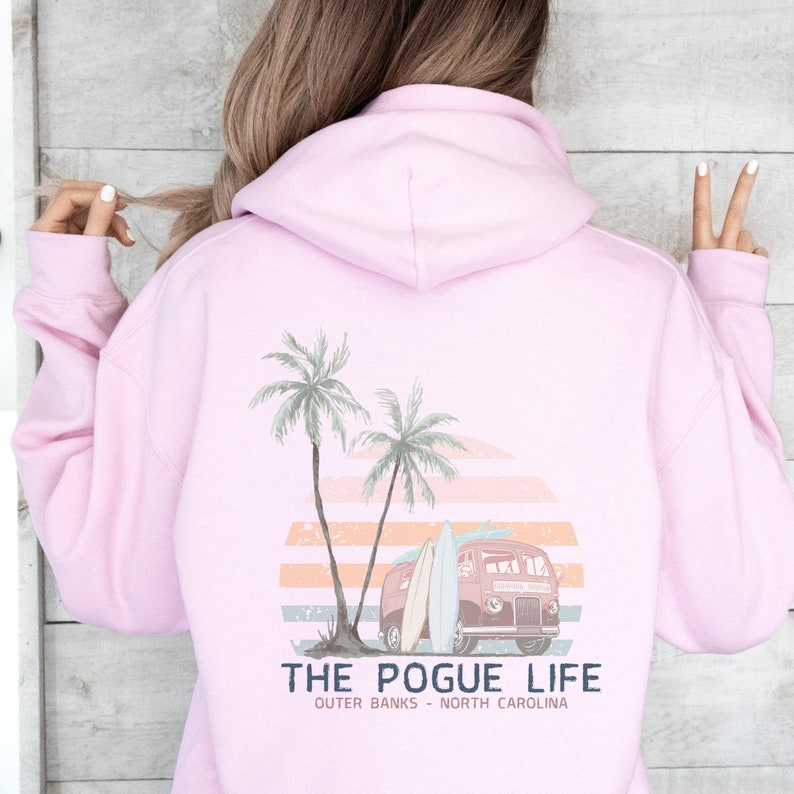 Op de afbeelding: Roze hoodie met een grafische print van een vintage busje met surfplanken, palmbomen en een zonsondergang. De tekst "THE POGUE LIFE" en "OUTER BANKS - NORTH CAROLINA" staat onder de print.
