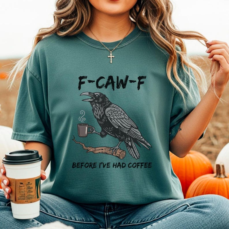 May include: Teal-colored t-shirt with a graphic of a raven holding a coffee cup, perched on a branch. The text "F-CAW-F" is above the raven, and "BEFORE I'VE HAD COFFEE" is below. The shirt is a casual, comfortable style.
