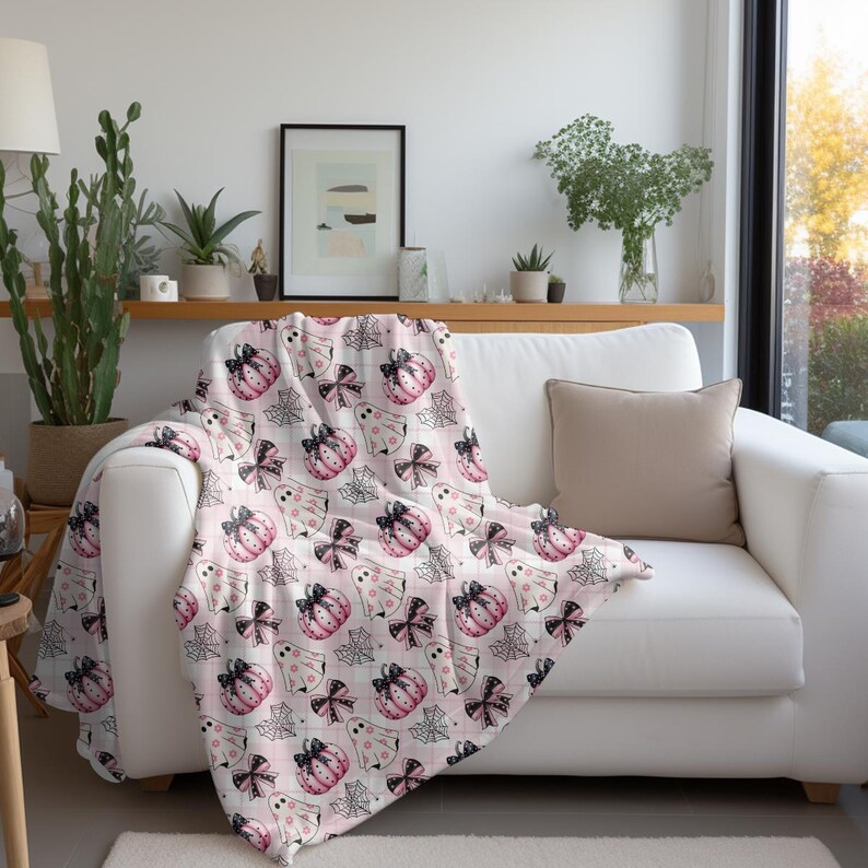 May include: A white throw blanket with a pink and black Halloween themed pattern. The pattern features pumpkins, ghosts, bows, and spiderwebs.