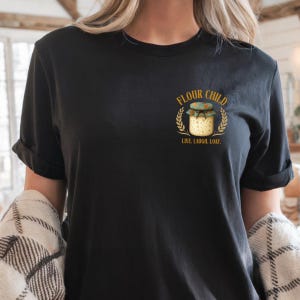 May include: Black t-shirt with a gold graphic of a jar with a floral lid, wheat stalks, and the words "FLOUR CHILD" and "LIVE. LAUGH. LOAF." The shirt has rolled-up sleeves.