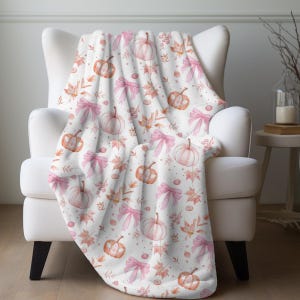 May include: A white throw blanket with a pink and orange watercolor pattern of pumpkins and bows.
