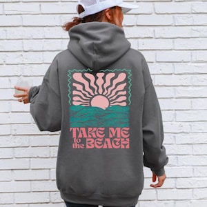 May include: Dark grey hoodie with a graphic on the back. The graphic shows a pink sun, teal waves, and the text "TAKE ME to the BEACH" in pink. The hoodie has a hood and long sleeves.