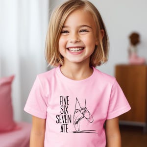 May include: A light pink t-shirt featuring a black graphic of ballet shoes and the words "FIVE SIX SEVEN ATE". The shirt is short-sleeved and worn by a smiling child.