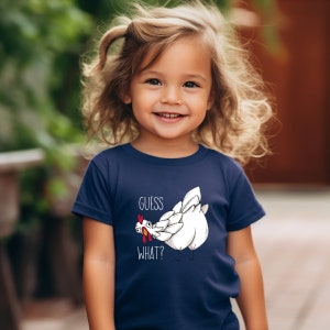 May include: A young child wearing a navy blue t-shirt with a white chicken illustration and the text "GUESS WHAT?" printed on it.