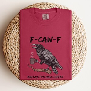 May include: A maroon t-shirt with the text "F-CAW-F" above a black and gray illustration of a raven holding a coffee cup. The text "BEFORE I'VE HAD COFFEE" is below the raven.