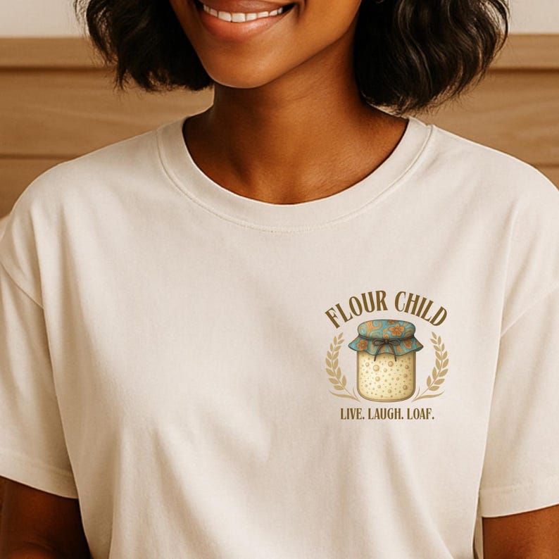 May include: A cream-coloured t-shirt with a graphic of a jar with a floral lid, wheat stalks, and the words "FLOUR CHILD" and "LIVE. LAUGH. LOAF." in a circular design. The shirt is made of a soft material.