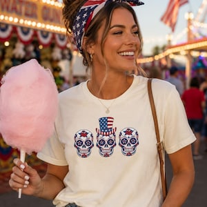 May include: A cream-coloured short-sleeve t-shirt featuring a graphic of three sugar skulls. The central skull wears a top hat with a red, white, and blue American flag pattern. The skulls are decorated with blue and red floral and star motifs.