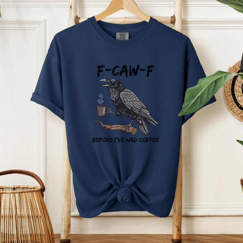 May include: Navy blue t-shirt featuring a graphic of a raven holding a coffee cup on a branch, with the text "F-CAW-F" above and "BEFORE I'VE HAD COFFEE" below. The raven is black and gray, and the coffee cup is brown.