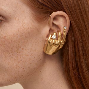 May include: A gold-colored ear cuff and stud earring set. The ear cuff is shaped like a hand holding a large, round, clear gemstone. A smaller gemstone is set in the earlobe. The jewelry is worn on a person's ear.