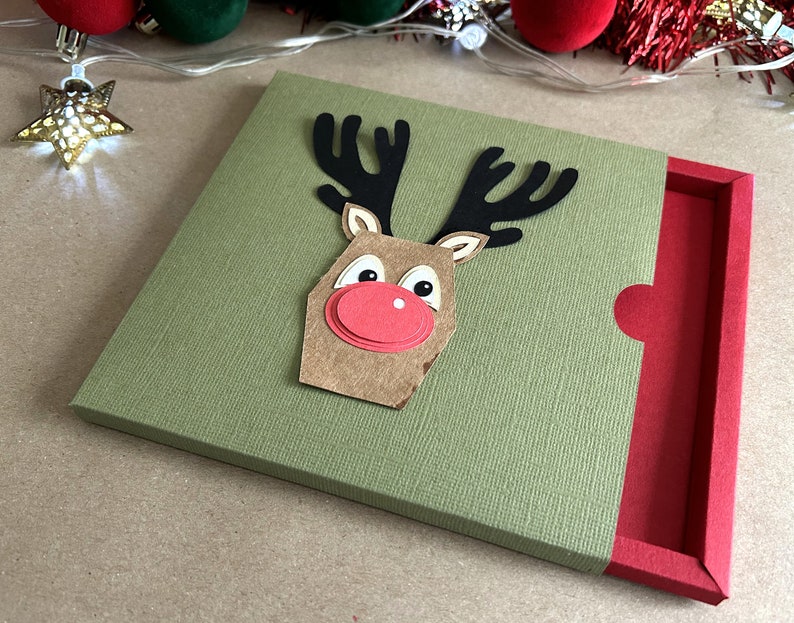 Puede incluir: Una caja de regalo verde con una cara de reno roja en la parte delantera. La caja tiene una solapa roja en el lateral.
