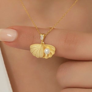 May include: A gold necklace with a delicate chain and a pendant shaped like a seashell with a pearl inside. The pendant is detailed with a textured surface.