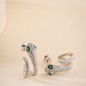 Puede incluir: Un par de pendientes plateados con forma de serpiente, con un engaste pavé de cristales transparentes a lo largo del cuerpo y la cabeza. Cada pendiente está acentuado con dos pequeñas piedras verdes en forma de lágrima como ojos y cierre de presión.