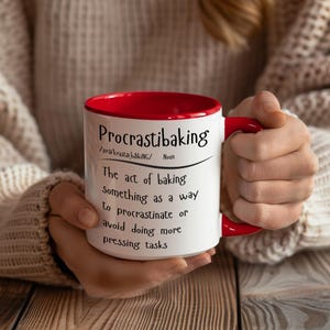 May include: White ceramic mug with a red rim. The mug has a black and white definition of the word "Procrastibaking" with the definition: "The act of baking something as a way to procrastinate or avoid doing more pressing tasks."
