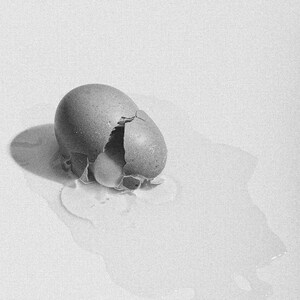 May include: A monochrome image of a cracked egg. The shell is broken, showing the yolk and white. The egg rests on a white surface, with the egg white spreading out. The lighting creates shadows.