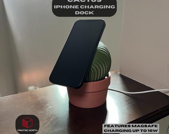 May include: A black iPhone is charging on a wireless charging dock shaped like a cactus in a terracotta pot. The dock features MagSafe charging up to 15W (USB-C).