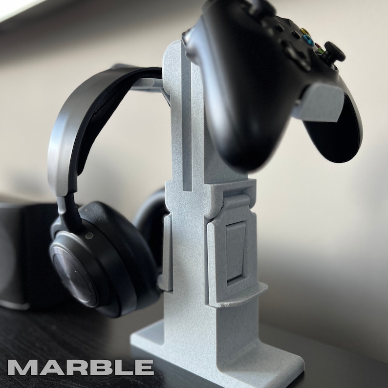 May include: A grey 3D printed stand with a black headset and a black video game controller. The stand has a slot for the headset and a groove for the controller. The stand is labelled "MARBLE".