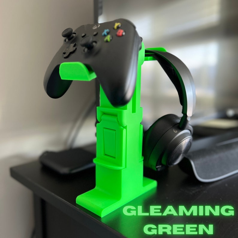 May include: A bright green 3D printed stand for a video game controller and headphones. The stand has a slot for the controller and a hook for the headphones. The stand is on a black surface. GLEAMING GREEN text is visible at the bottom of the image.