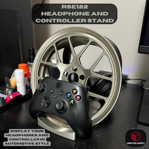 Può includere: Un supporto per cuffie e controller argentato con la scritta "RSE122 HEADPHONE AND CONTROLLER STAND". Un controller nero poggia sul supporto, con un supporto per cuffie a forma di ruota argentata dietro. È visibile anche la scritta "DISPLAY YOUR HEADPHONES AND CONTROLLER IN AUTOMOTIVE STYLE".