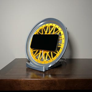 Puede incluir: Un soporte para teléfono gris y amarillo con forma de rueda de coche. Un teléfono inteligente negro está en el soporte.