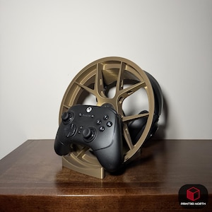 Op de afbeelding: Een goudkleurige, wielvormige standaard voor gamingheadsets en controllers. Een zwarte Xbox-controller rust op de standaard. De standaard is gemaakt van goudkleurig materiaal en staat op een houten oppervlak.