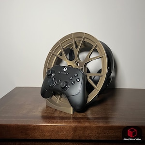 Könnte beinhalten: Ein goldfarbener, 3D-gedruckter Gaming-Headset- und Controller-Ständer. Der Ständer hat die Form eines Autorads und hält einen schwarzen Xbox-Controller und ein Headset. Der Ständer steht auf einer dunkelbraunen Holzoberfläche.