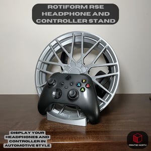 May include: A silver and black 3D printed stand shaped like a car wheel, designed to hold a gaming controller and headphones. The stand features the text "ROTIFORM RSE HEADPHONE AND CONTROLLER STAND" and "DISPLAY YOUR HEADPHONES AND CONTROLLER IN AUTOMOTIVE STYLE".